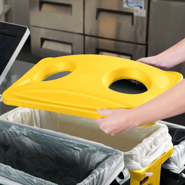A person holding a yellow Rubbermaid Slim Jim recycling container lid over a trash can.