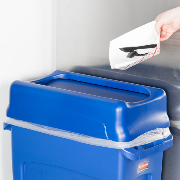 A hand putting a piece of paper into a blue Rubbermaid Slim Jim trash can lid.