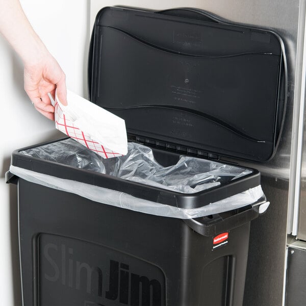A hand putting a paper bag in a black Rubbermaid Slim Jim trash can.