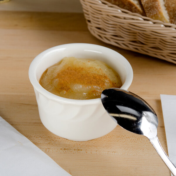 A white Tuxton china bouillon cup filled with brown food and a spoon on a table with a basket of bread.