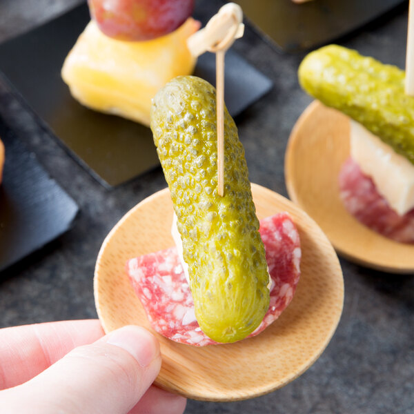 A hand holding a pickle on a toothpick over a small plate of food.