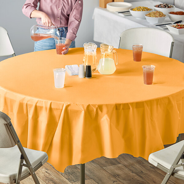 A table set with a pumpkin spice orange round plastic table cover and glasses of orange liquid on an outdoor table.