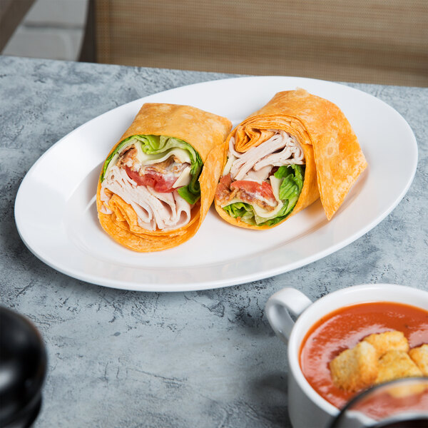 A white Elite Global Solutions oval melamine plate with a sandwich and tortilla wrap on it.