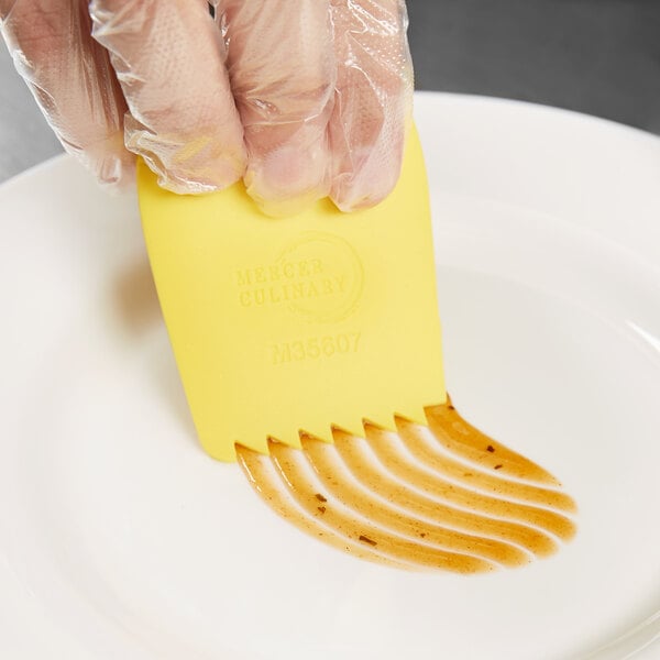 A person's gloved hand using a Mercer Culinary yellow silicone wedge plating tool over a white plate.