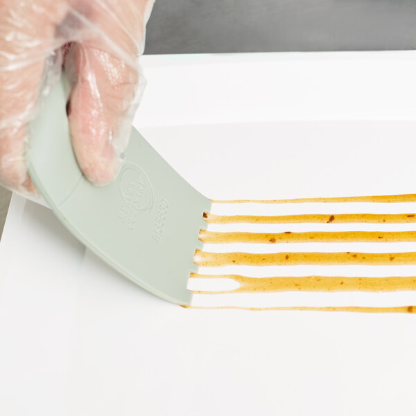 A hand using a Mercer Culinary silicone wedge plating tool to spread brown liquid on a white surface.