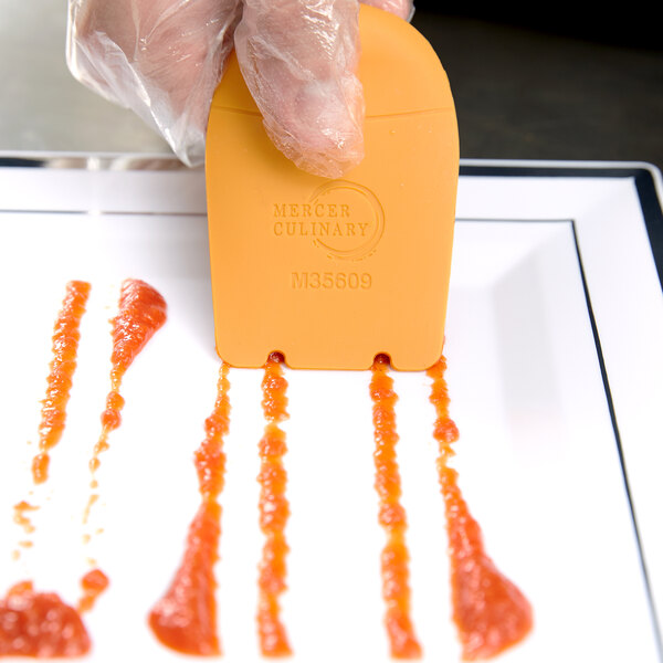 A person using a Mercer Culinary horseshoe arch plating tool to place a piece of orange food on a plate.