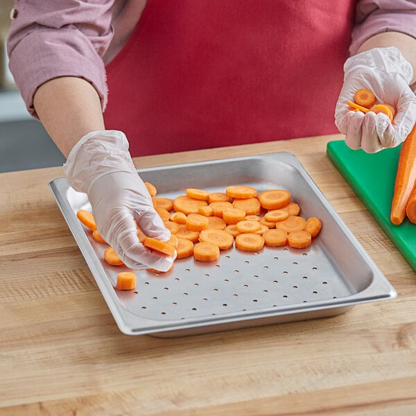 A person wearing gloves and holding a Choice 1/2 size stainless steel tray of carrots cuts a carrot.