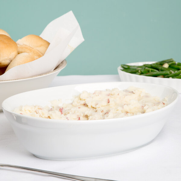 A white oval serving bowl filled with mashed potatoes next to a plate of green beans.