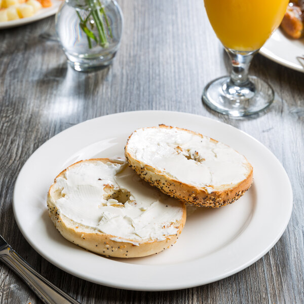 A Libbey white porcelain plate with a bagel with cream cheese on it next to a glass of orange juice.