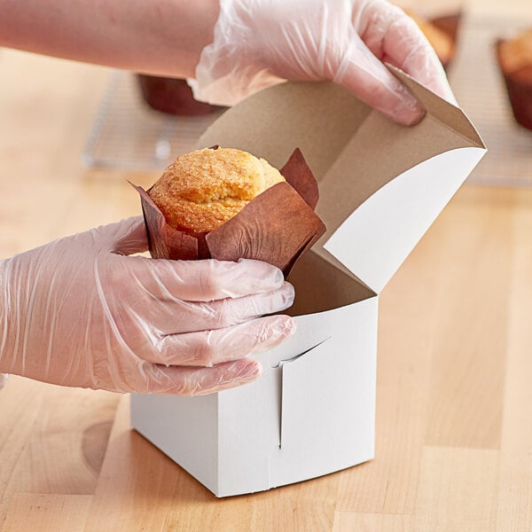 A person in gloves holding a cupcake in a white bakery box.