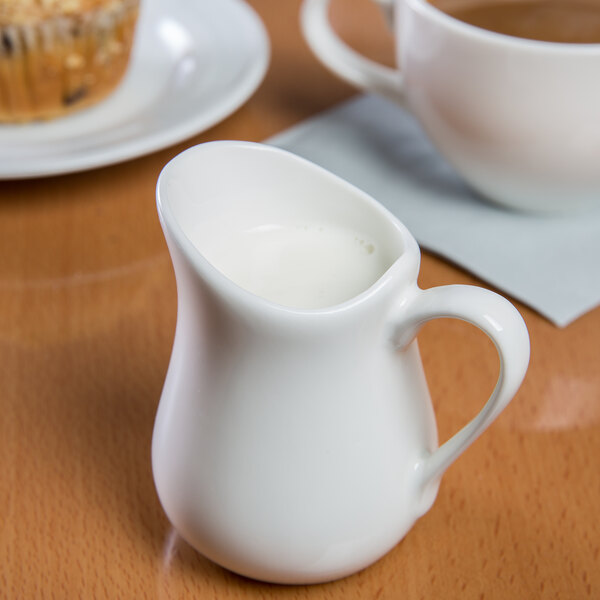 A white Libbey porcelain creamer with a handle on a table with a cup of coffee.