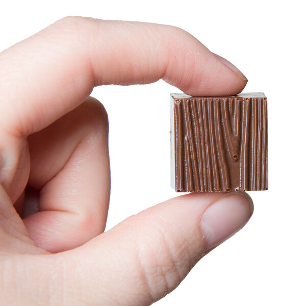 A hand holding a small square chocolate with a wood texture.
