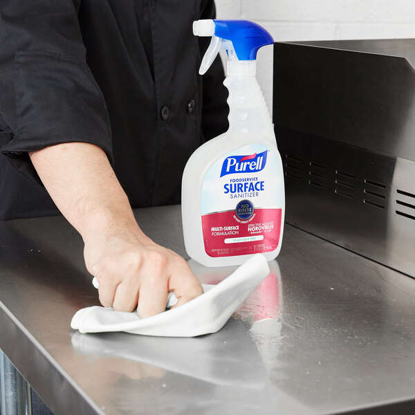 A hand wiping a stainless steel counter with a cloth using Purell Fragrance Free Foodservice Surface Sanitizer.