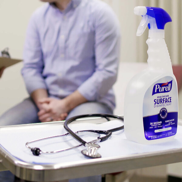 A man sitting at a table with a Purell Healthcare Surface Disinfectant spray bottle next to a stethoscope.