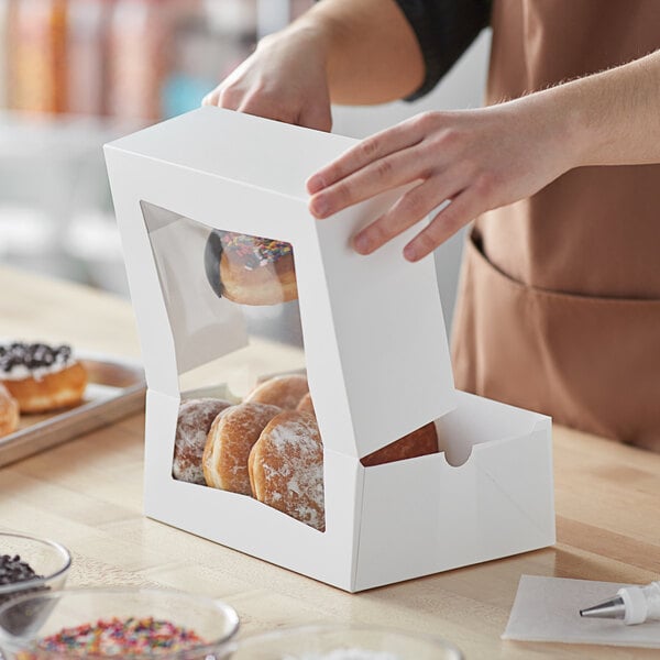 A person opening a Baker's Mark white bakery box to reveal doughnuts inside.