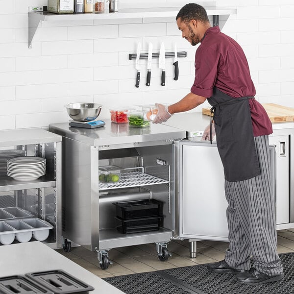 A man in a chef's uniform putting food into an Avantco undercounter refrigerator in a professional kitchen.