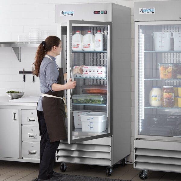 A woman opening an Avantco stainless steel glass door reach-in refrigerator in a professional kitchen.