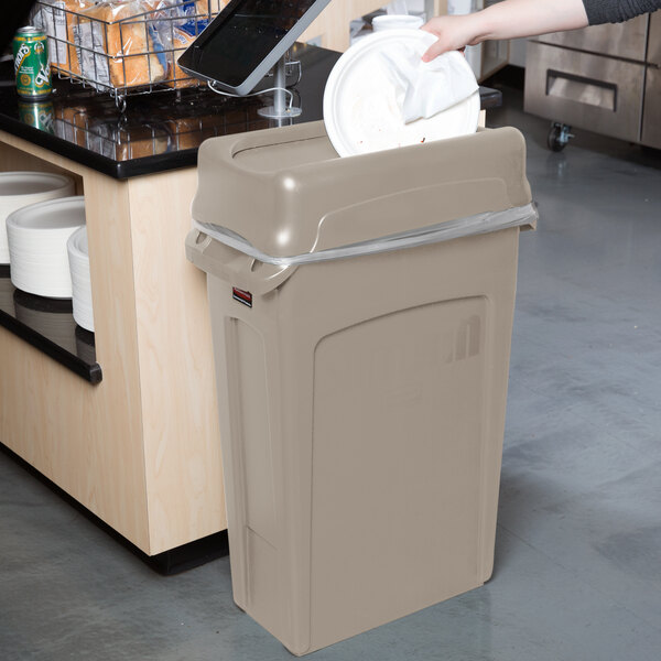 A person throwing a white plate into a tan Rubbermaid Slim Jim rectangular trash can.