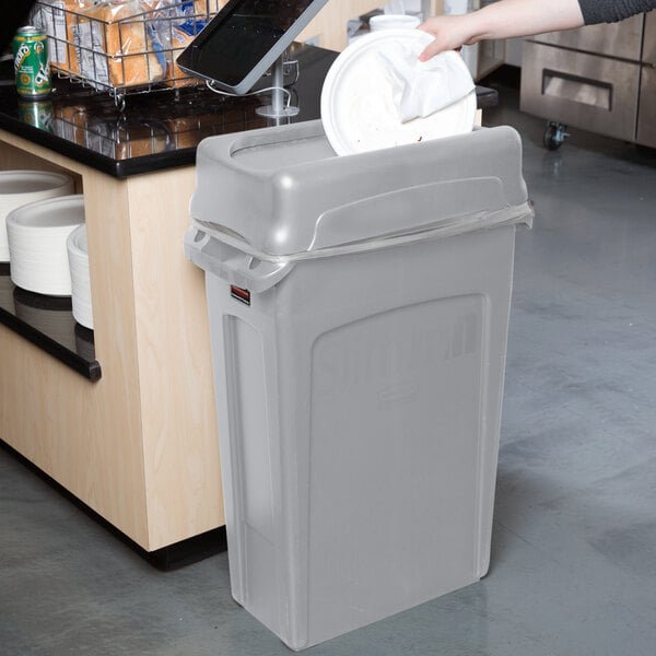 A person throwing a white paper plate into a Rubbermaid Slim Jim trash can with a gray lid.