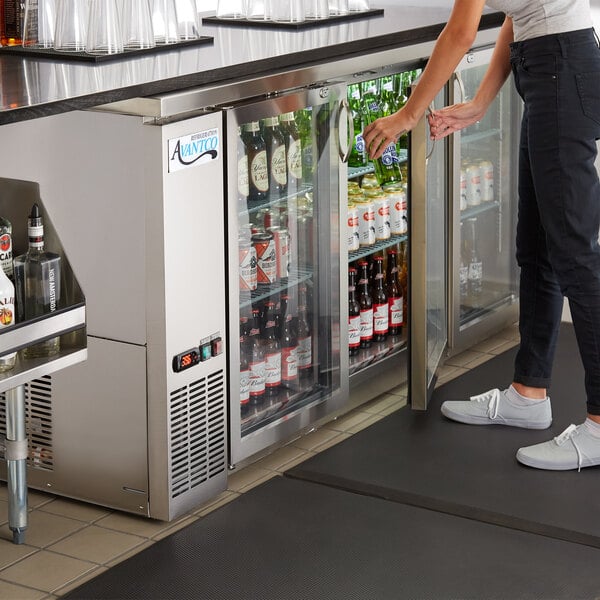 A woman standing next to an Avantco stainless steel back bar refrigerator with glass doors.