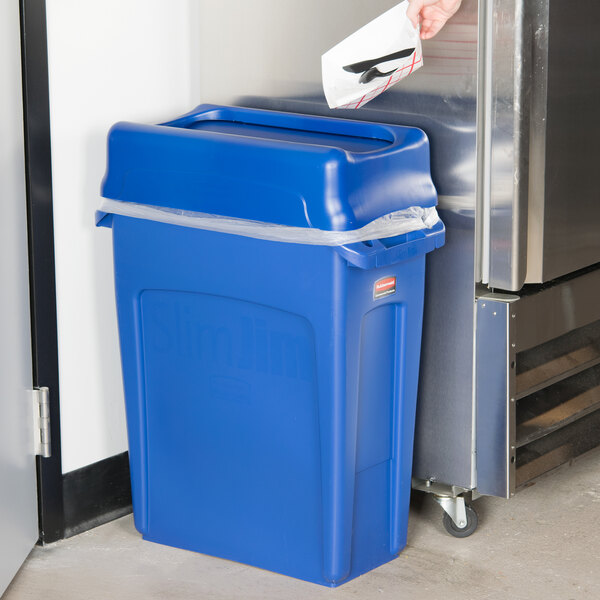 A person holding a blue Rubbermaid Slim Jim recycling bin next to a stainless steel refrigerator.
