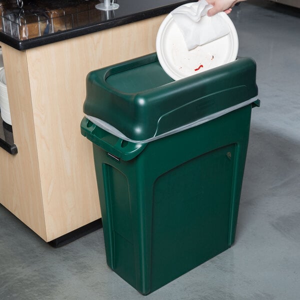 A hand putting a white plate into a green Rubbermaid Slim Jim trash can
