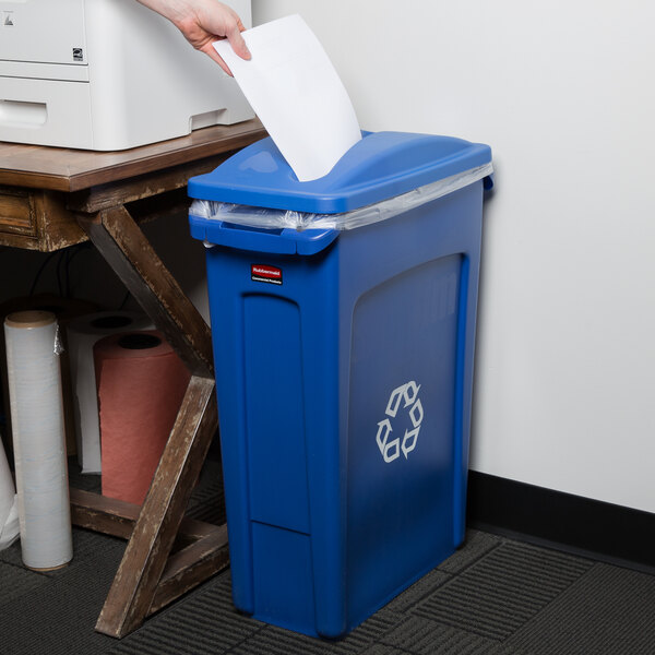 A hand putting paper into a blue Rubbermaid recycling bin.