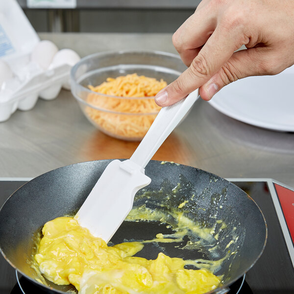 A hand using a white Rubbermaid high temperature silicone spatula to stir eggs in a pan.