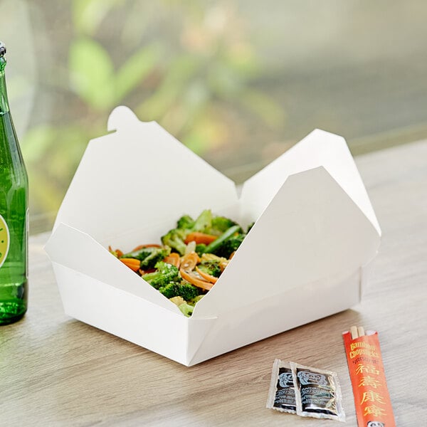 A white Choice folded paper take-out box of food with broccoli and a bottle on a table.