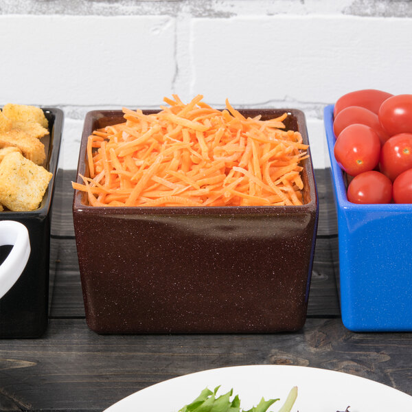 A Tablecraft maroon speckle bowl filled with shredded carrots on a table with a blue container of tomatoes.