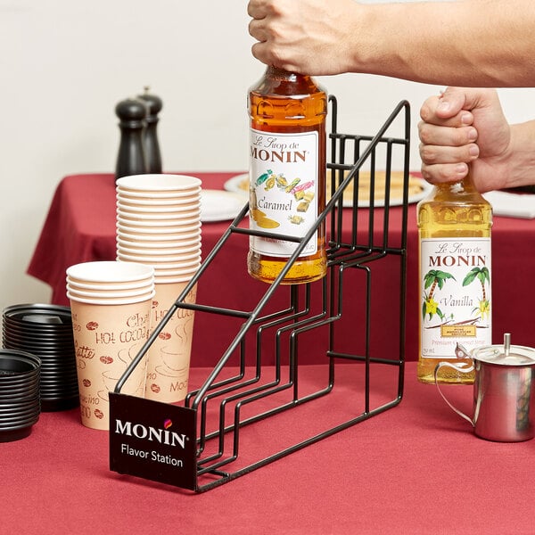 A person holding a Monin syrup bottle over a metal cup on a Monin syrup rack.