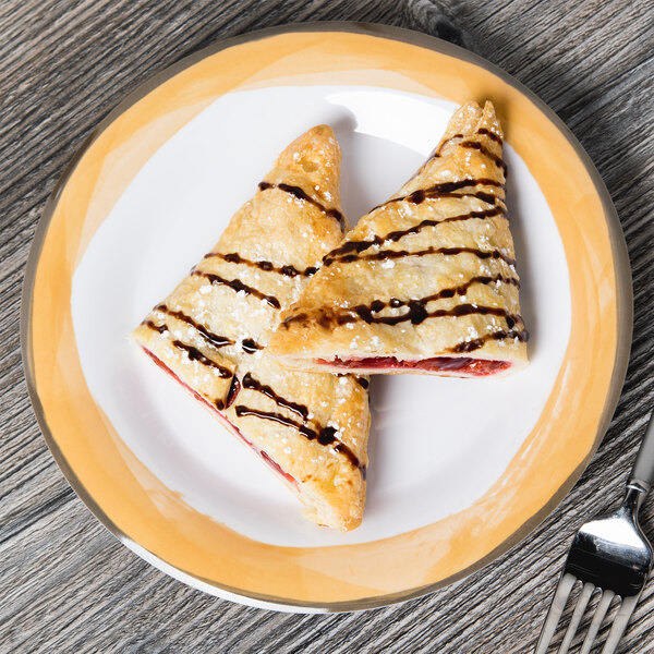 A Kanello white melamine plate with a yellow edge holding two pastries with chocolate and strawberry sauce on it.