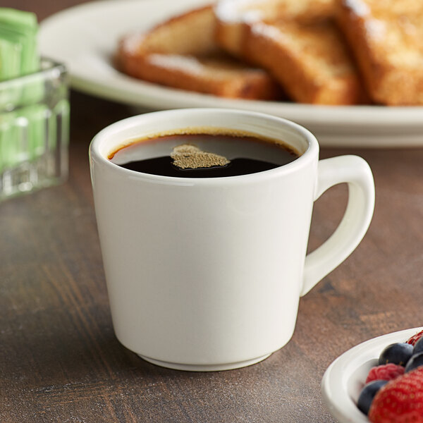 An Acopa tall ivory stoneware cup filled with brown liquid sits on a table next to a plate of toast.