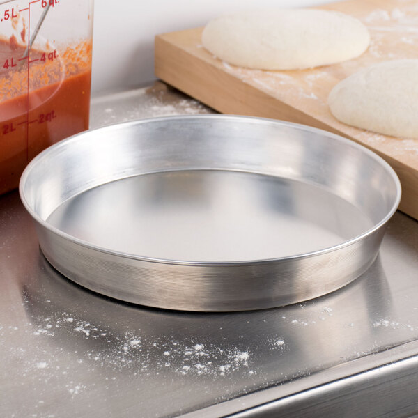 An American Metalcraft aluminum pizza pan on a counter with dough on it.