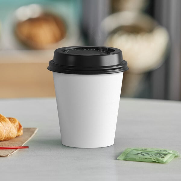 A white Choice paper hot cup with a black lid on a table with a croissant.