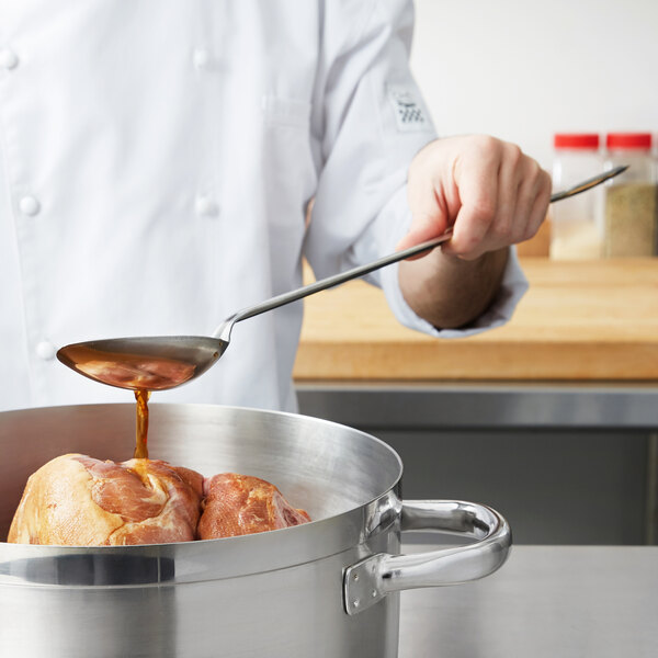 A person using a Vollrath solid stainless steel basting spoon to pour sauce over meat in a pot.