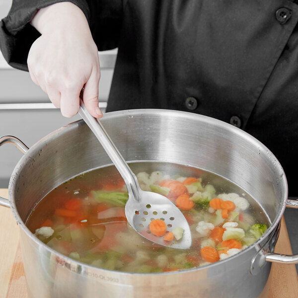 A person using a Vollrath perforated stainless steel basting spoon to stir soup in a pot.