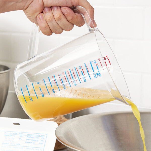 A hand pouring orange juice into a Rubbermaid clear polycarbonate measuring cup.