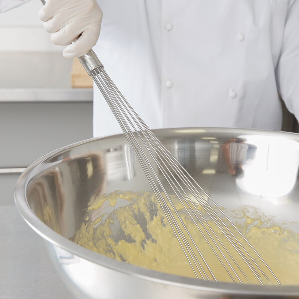 A person in a white coat using a Vollrath stainless steel whisk to mix yellow liquid in a bowl.