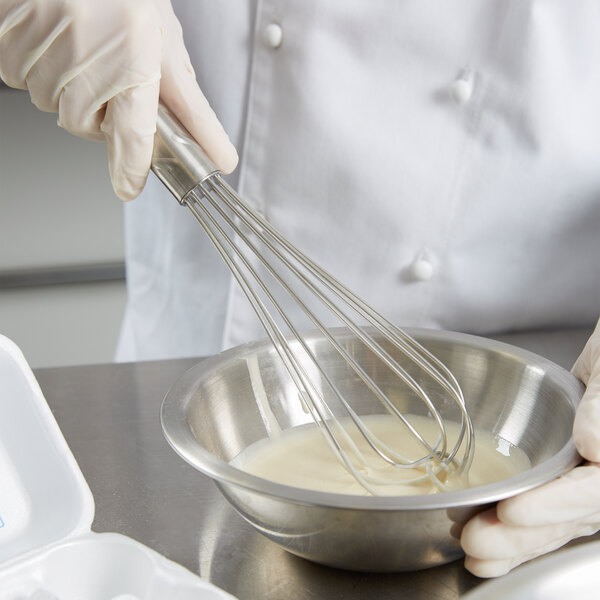 A person in a white coat using a Vollrath stainless steel whisk to mix ingredients in a bowl.