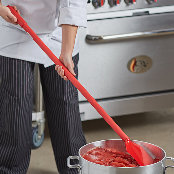 A person using a Carlisle Sparta paddle with a red handle to stir a pot of red sauce.