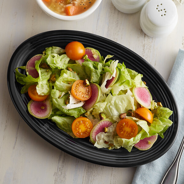 A Tuxton black oval china platter with salad including tomatoes and radishes.