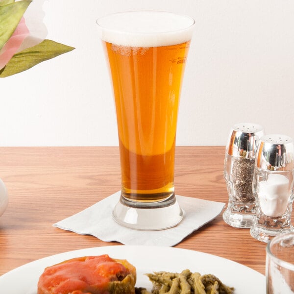 A Libbey pilsner glass filled with beer next to a plate of food on a table.