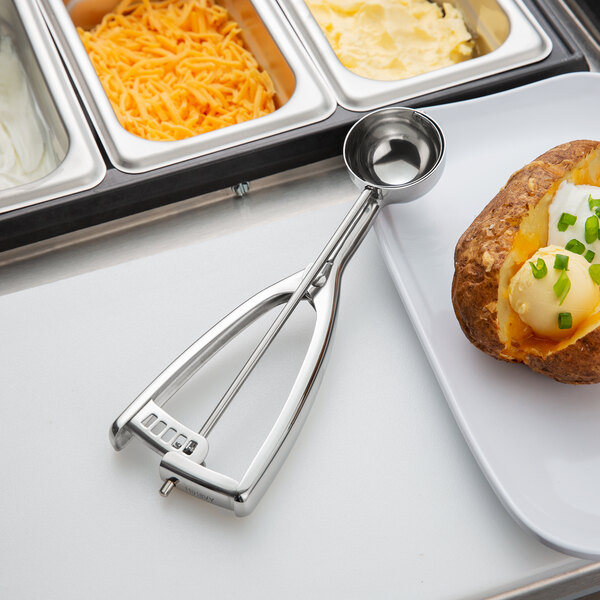 A baked potato with cheese and green onions with a Vollrath stainless steel scoop on a plate.