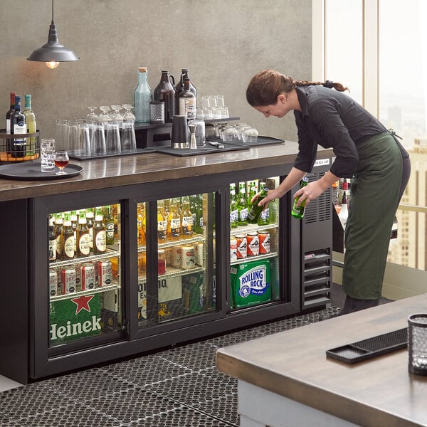 A woman putting a bottle of beer in a Beverage-Air black sliding glass door back bar refrigerator.