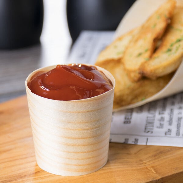 A Tablecraft mini wooden serving cup filled with ketchup on a wood table with french fries.