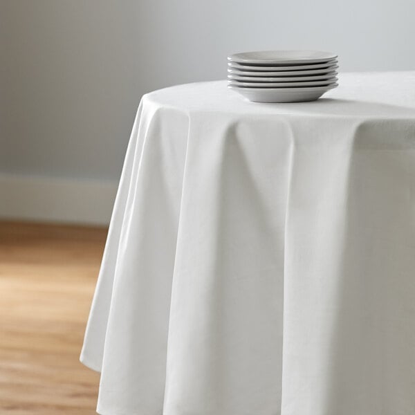 A white Intedge round tablecloth on a table with a stack of plates.