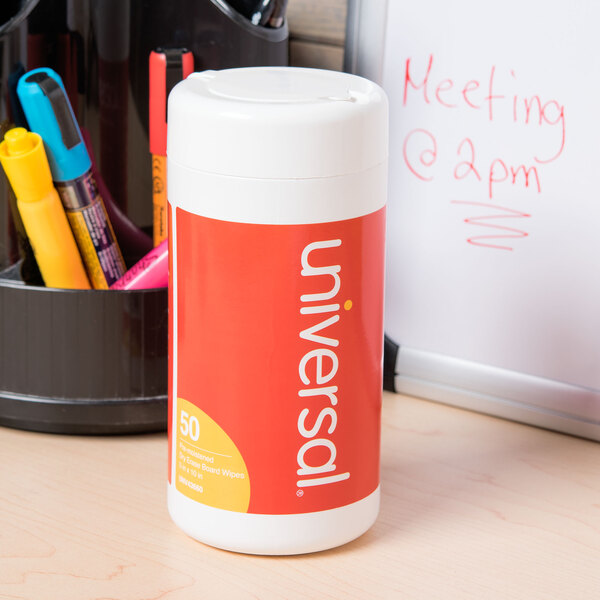A white container with a red label for Universal Pop-Up Dry Erase Cleaning Wipes on a desk.