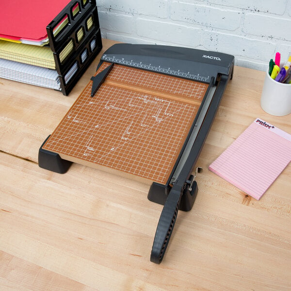 An X-Acto paper cutter on a wood table with a stack of paper.