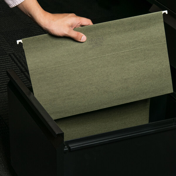 A hand places a green UNV14151 Legal Size Box Bottom Hanging File Folder in a file cabinet.